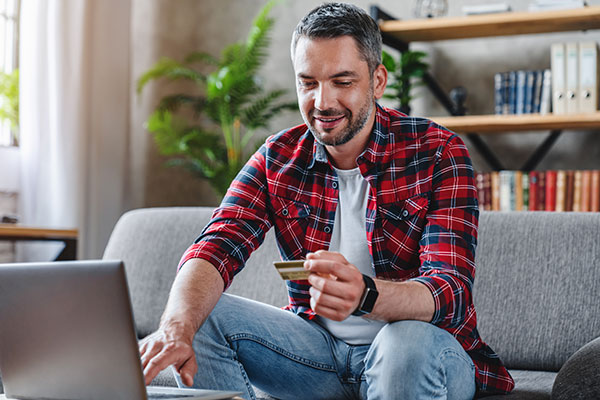 A man is wearing a red flannel shirt sitting in living room, holding a credit card and ordering something online with his laptop.