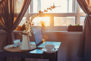 A table with a laptop, a cup of coffee, flowers in a vase, and a candle in front of a window with the sun is showing through, representing working from home.